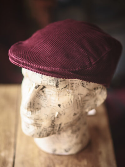 A model head wearing a traditional red corduroy flat cap